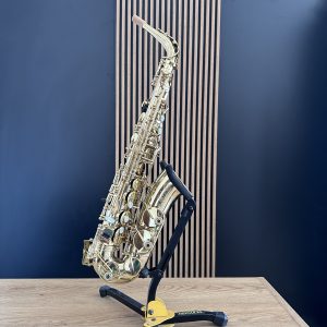 Gold saxophone resting in a black-yellow stand on a light wooden table against a dark blue wall with vertical slats behind it.