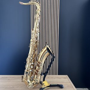 Gold saxophone resting on a black stand on a light wooden table against a blue wall, with the curved neck and keys visible.